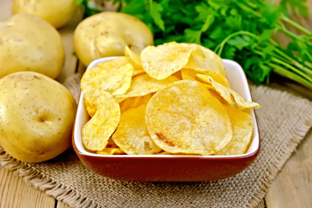 Potato chips in a brown and white bowl, with whole potatoes in the background.