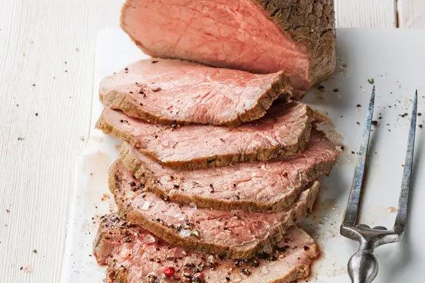 Roast Beef half sliced on a white cutting board with a fork next to it, on a wooded background.