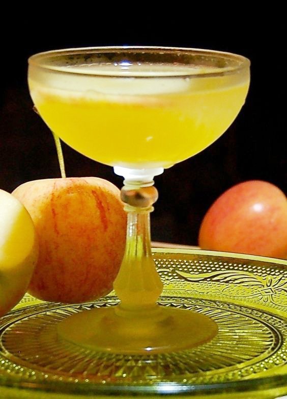 Closeup of an Apple Cider Vodka Martini in a clear glass on top of a glass plate surrounded by apples, with a black background.