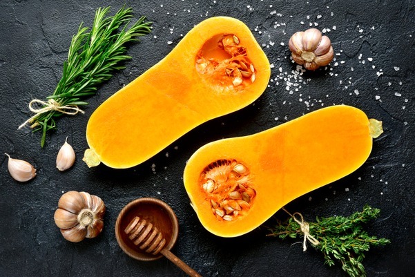 Butternut squash cut in half on a black background surrounded by herbs.