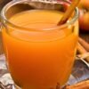 A clear glass mug filled with hot apple cider sits on a saucer, garnished with a cinnamon stick. A star anise pod and cinnamon stick rest on the table in front, with fresh apples and a basket softly blurred in the background, giving the scene a warm autumn feel.