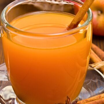 A clear glass mug filled with hot apple cider sits on a saucer, garnished with a cinnamon stick. A star anise pod and cinnamon stick rest on the table in front, with fresh apples and a basket softly blurred in the background, giving the scene a warm autumn feel.