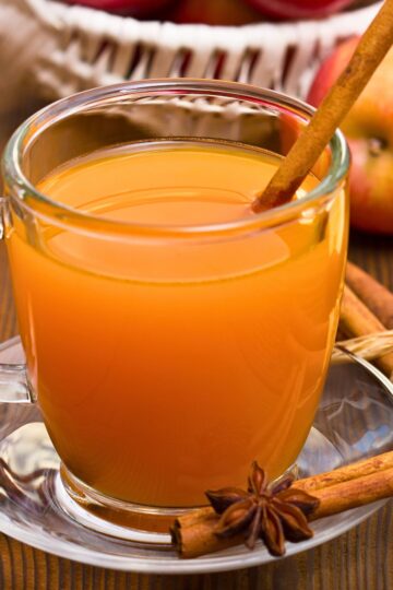 A clear glass mug filled with hot apple cider sits on a saucer, garnished with a cinnamon stick. A star anise pod and cinnamon stick rest on the table in front, with fresh apples and a basket softly blurred in the background, giving the scene a warm autumn feel.