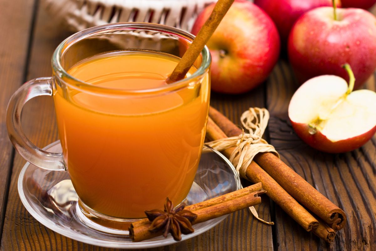 A glass mug of hot apple cider sits on a clear saucer, garnished with a cinnamon stick. Star anise and bundled cinnamon sticks rest nearby on a rustic wooden table. Fresh red apples, including a sliced one, are in the background, adding a cozy fall harvest feel.