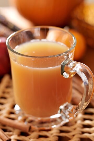 Clear glass mug of warm apple cider on a woven placemat, steam rising with cinnamon sticks beside it. Soft-focus pumpkin, red apple, and decorative corn in the background—cozy fall drink, apple cider recipe, cinnamon, autumn kitchen.
