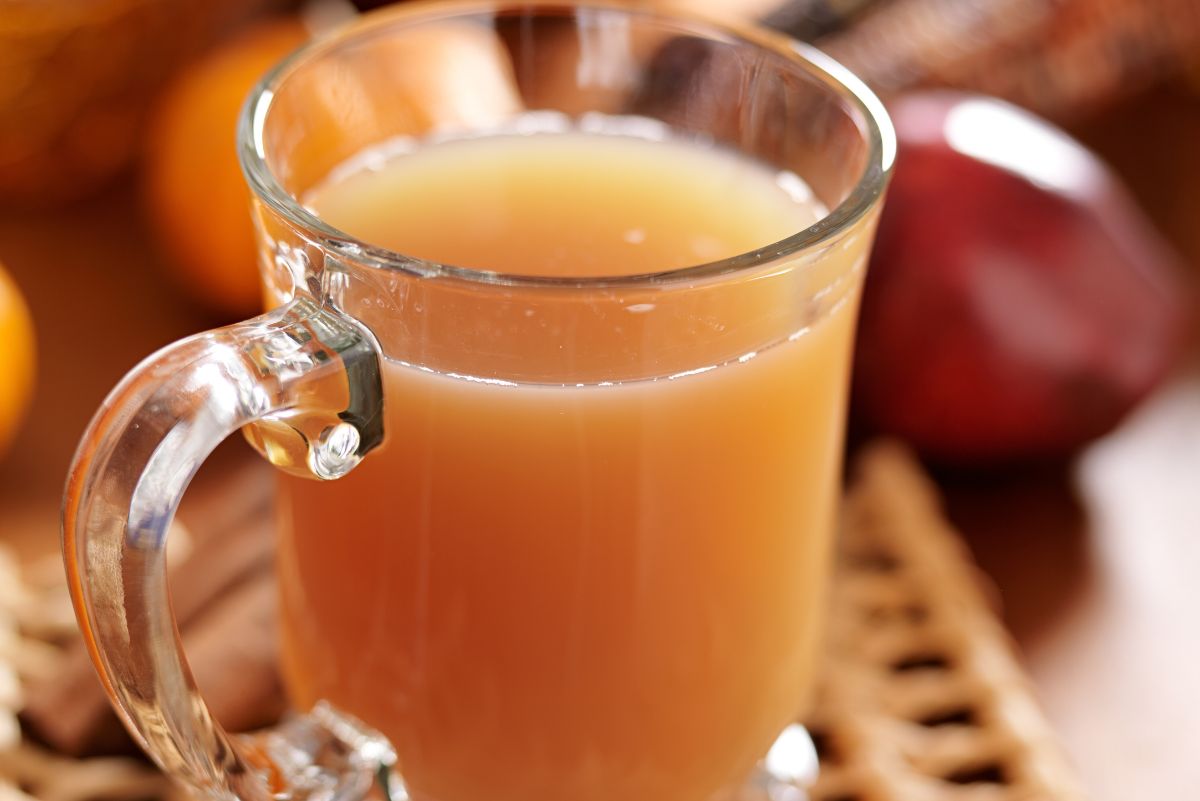 Close-up of a clear glass mug filled with warm apple cider on a woven placemat; soft-focus red apple and fall decor in the background. Cozy autumn drink, apple cider recipe, fall kitchen.