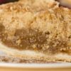Close-up of a Granny Smith apple crumb pie slice with buttery streusel topping, gooey cinnamon-spiced apple filling, and flaky pie crust on a white plate lightly dusted with cinnamon.