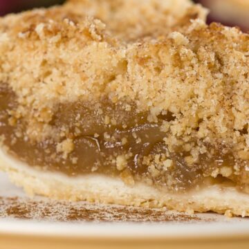 Close-up of a Granny Smith apple crumb pie slice with buttery streusel topping, gooey cinnamon-spiced apple filling, and flaky pie crust on a white plate lightly dusted with cinnamon.