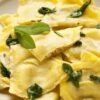 Homemade butternut squash ravioli served on a white plate, topped with fresh sage leaves and lightly coated in butter sauce. The golden pasta squares are tender with a rustic, handmade look, perfect for a cozy Italian-inspired meal.