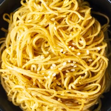 Asian sesame garlic noodles piled in a black bowl, with glossy spaghetti coated in a light golden sauce and sprinkled with white sesame seeds. Close-up food photo with a simple noodle dish on a dark stone background.