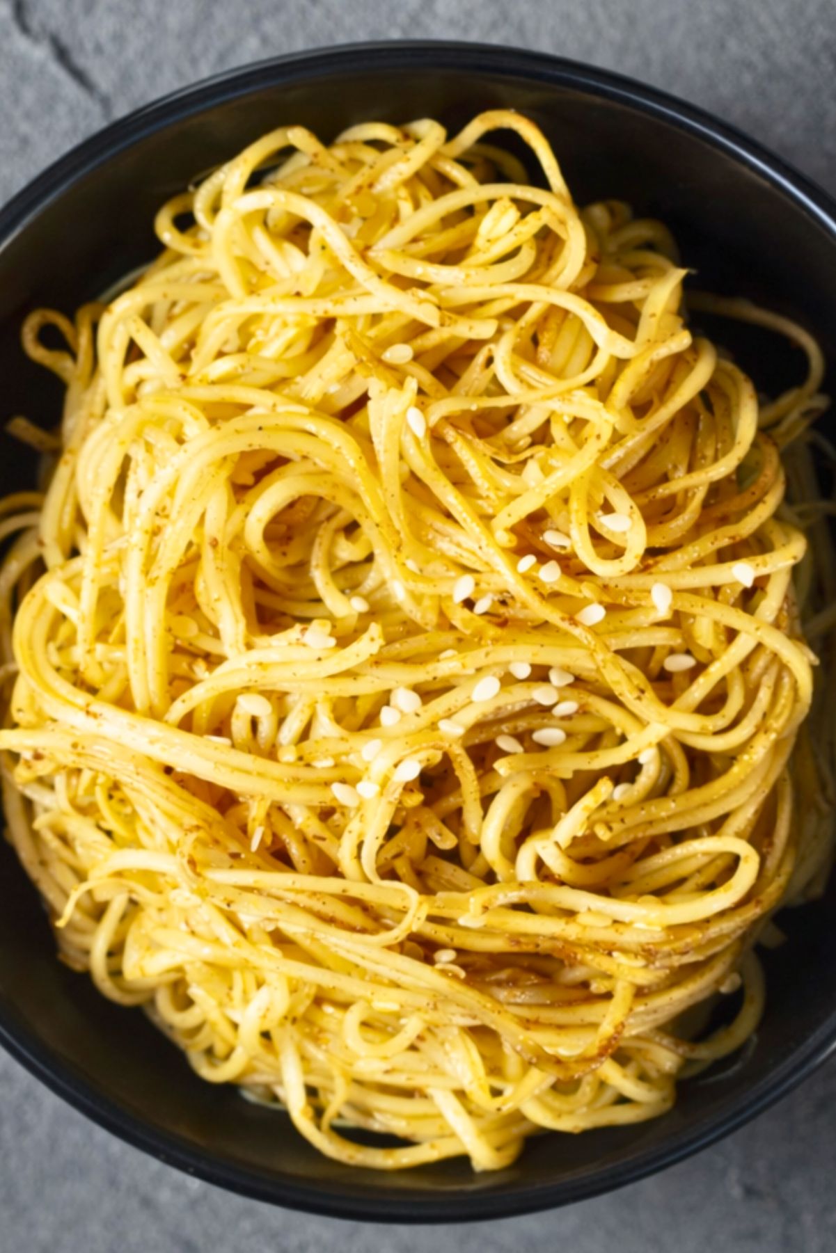 Asian sesame garlic noodles piled in a black bowl, with glossy spaghetti coated in a light golden sauce and sprinkled with white sesame seeds. Close-up food photo with a simple noodle dish on a dark stone background.