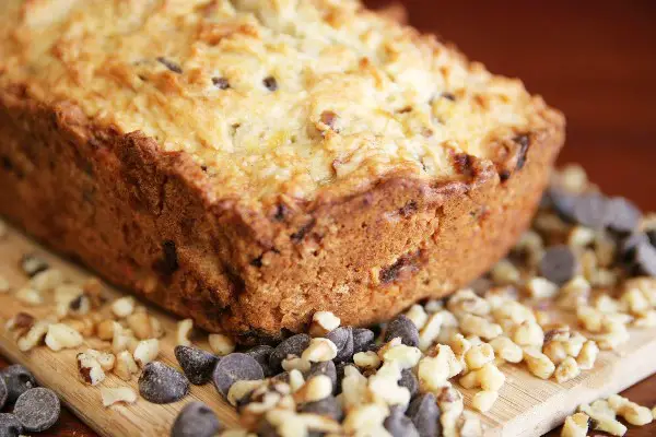 Zucchini bread on a cutting board, surrounded by chocolate chips and chopped walnuts.