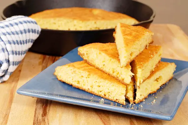 A pile of Cornbread slices in front of a cast iron skillet.