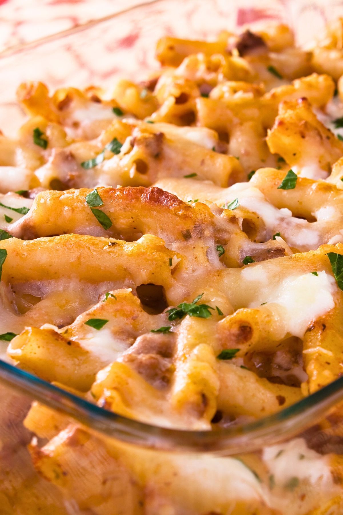 Close-up of a baked Italian macaroni and cheese casserole in a glass dish, with rigatoni pasta coated in creamy melted cheese, browned spots on top, bits of ground meat, and a sprinkle of fresh parsley, classic comfort food pasta bake for dinner.