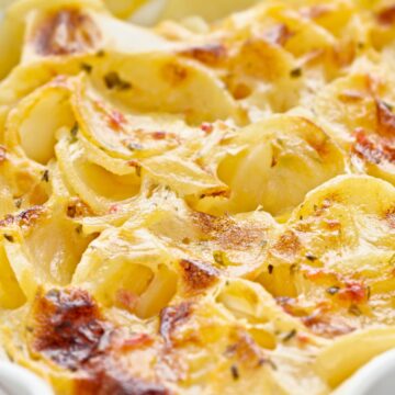 Close-up of cheesy au gratin scalloped potatoes with thinly sliced potatoes layered in a creamy cheese sauce. Golden browned spots and lightly crisp edges on top, baked in a white ceramic casserole dish. Rich, creamy potato side dish with melted cheese and herbs.