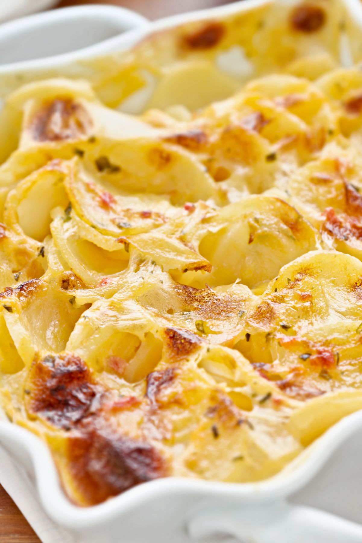 Close-up of cheesy au gratin scalloped potatoes with thinly sliced potatoes layered in a creamy cheese sauce. Golden browned spots and lightly crisp edges on top, baked in a white ceramic casserole dish. Rich, creamy potato side dish with melted cheese and herbs.