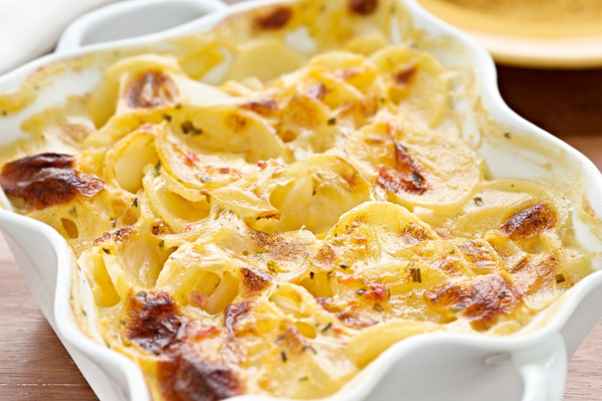 Au gratin scalloped potatoes baked in a white ceramic casserole dish, with thinly sliced potatoes layered in a creamy cheese sauce. Golden brown spots and lightly crisp edges on top, showing melted cheese, tender potatoes, and a rich, homemade potato casserole side dish.