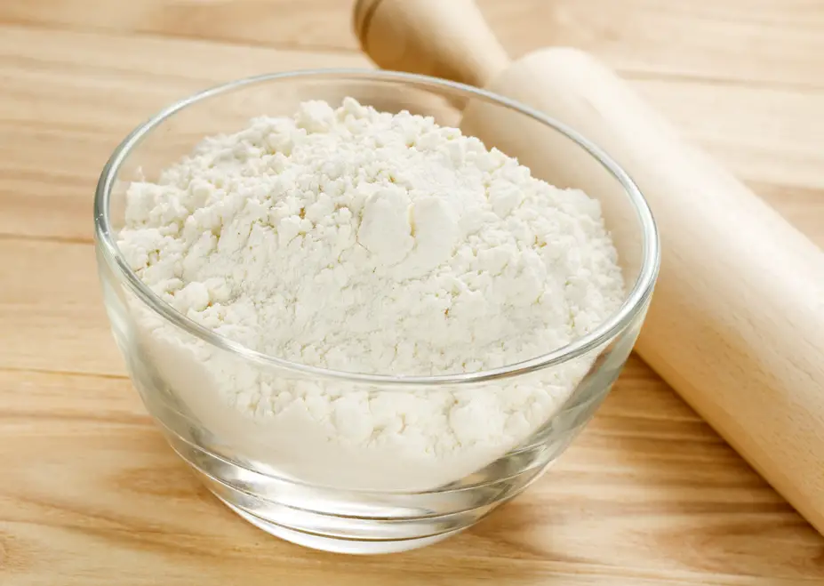 Flour in a clear glass bowl with a rolling pin next to it.