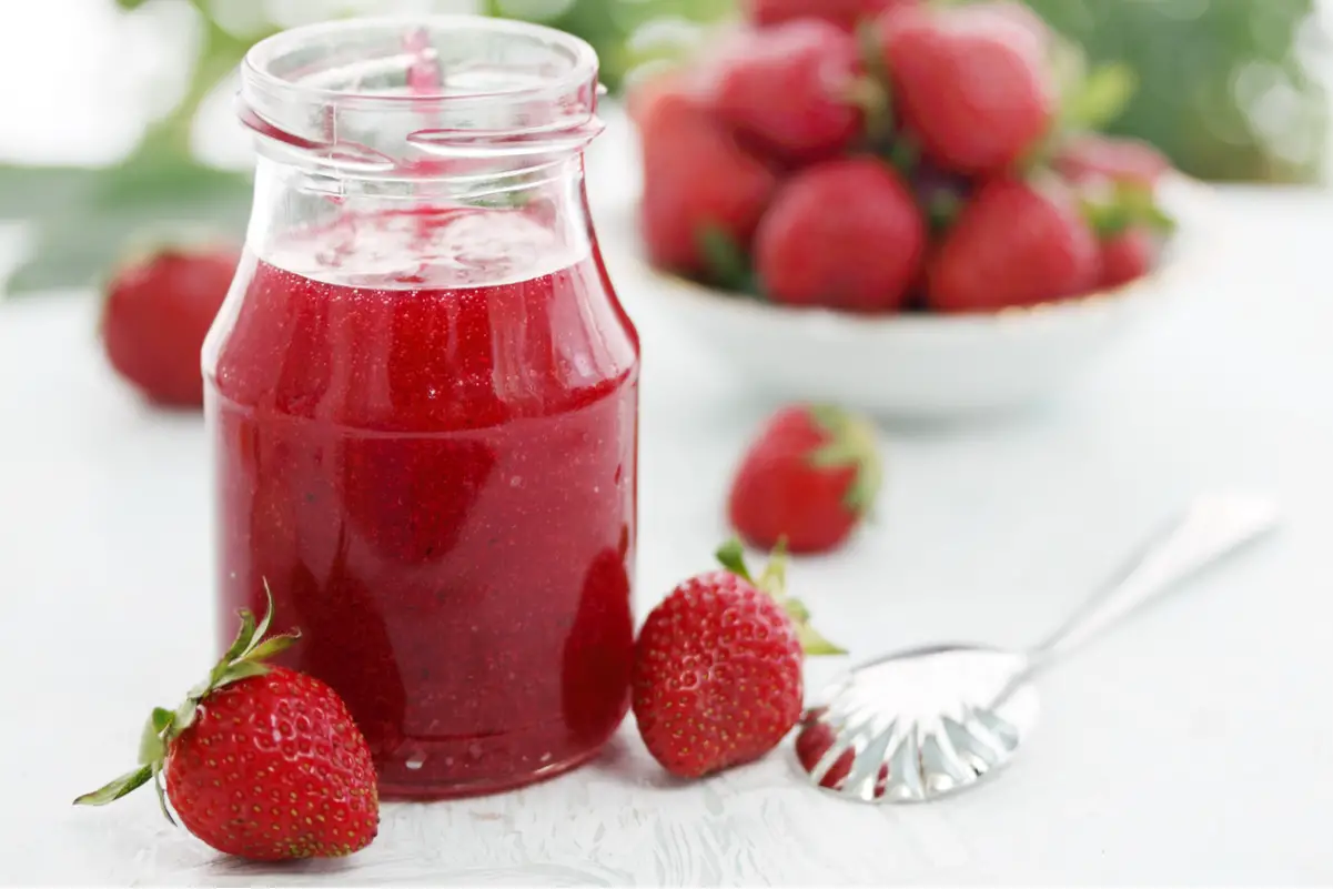 Easy Homemade Strawberry Sauce in a clear glass jar with strawberries in the background.
