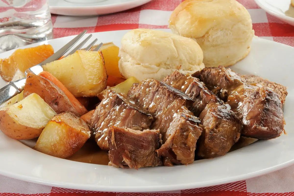 Easy Slow Cooker Pot Roast on a white dinner plate with carrots, potatoes, and a dinner roll. The plate is on a red and white checked tablecloth.