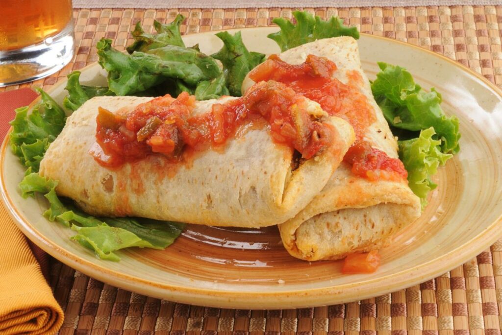 2 Mexican beef and bean chimichangas on a tan plate with an orange cloth napkin next to it.