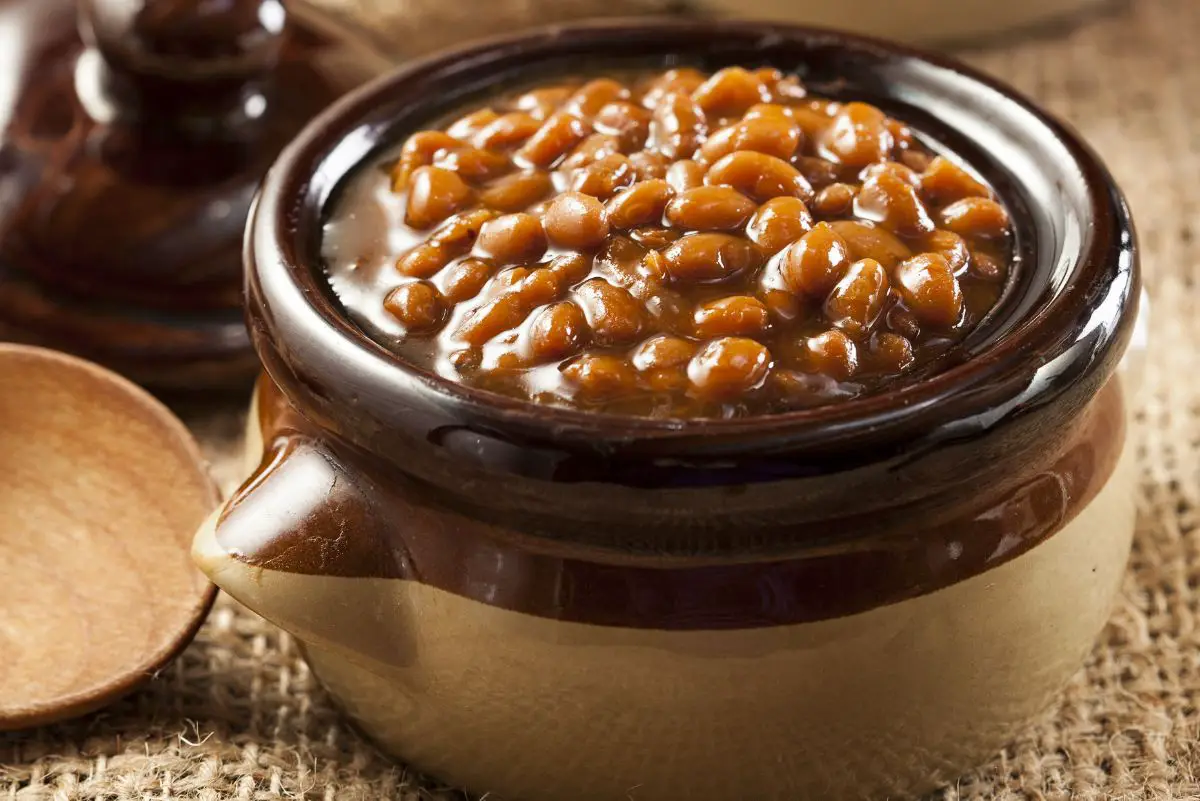 Homemade Boston Baked Beans in a brown crock with the lid laying on the table.