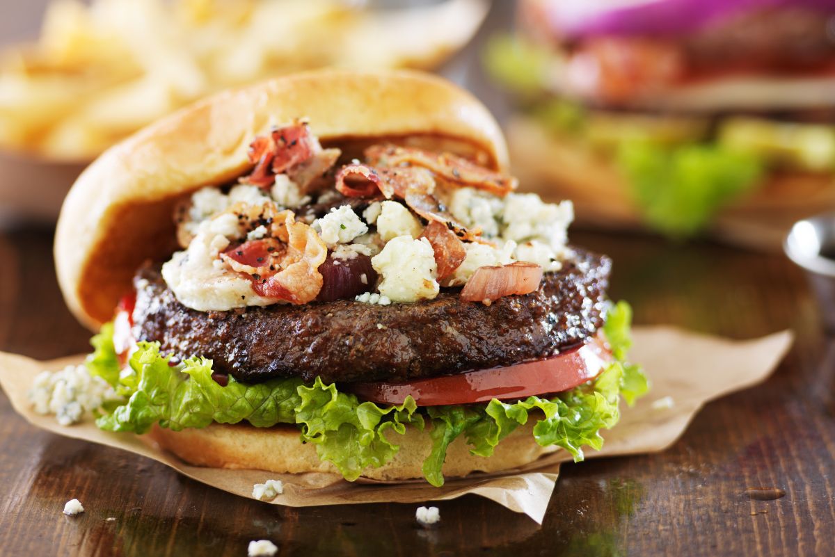 A Bacon Cheeseburgers with Blue Cheese on a dark wooden table with out of focus burgers behind it.