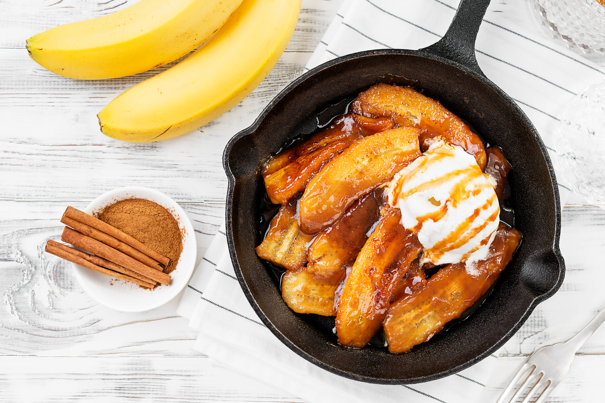 Overhead view of bananas foster in a cast iron skillet with a whole banana, and a small dish of cinnamon next to it.