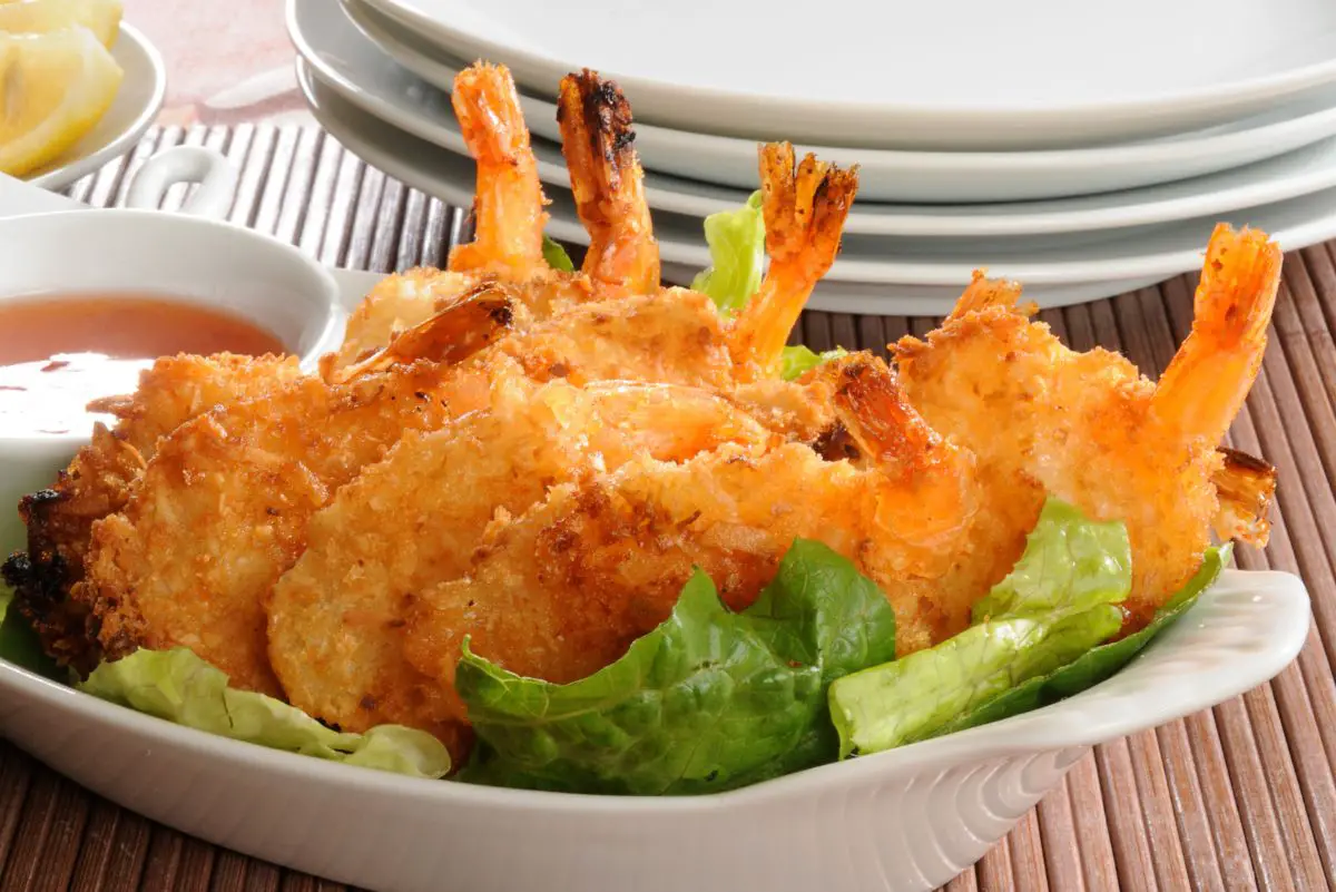 Crispy Coconut Shrimp in Beer Batter Recipe in a white bowl with a stack of white plates behind it.