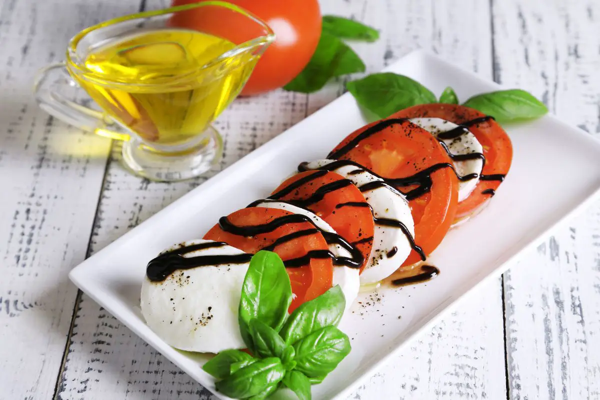 Overhead view of Italian Balsamic Caprese Salad on a rectangular, white plate with a tomato and bottle of oil next to it.