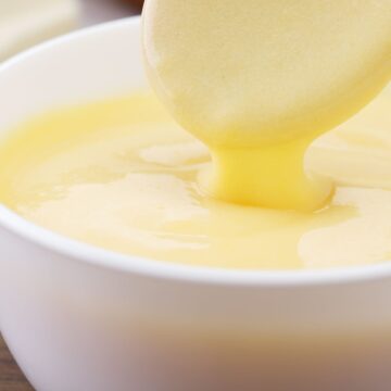 Closeup of Quick and Easy Hollandaise Sauce in a white bowl with a wooden spoon stirring the sauce.