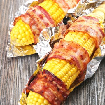 2 Bacon Wrapped Corn on the Cobs on a rustic surface.