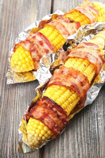 2 Bacon Wrapped Corn on the Cobs on a rustic surface.