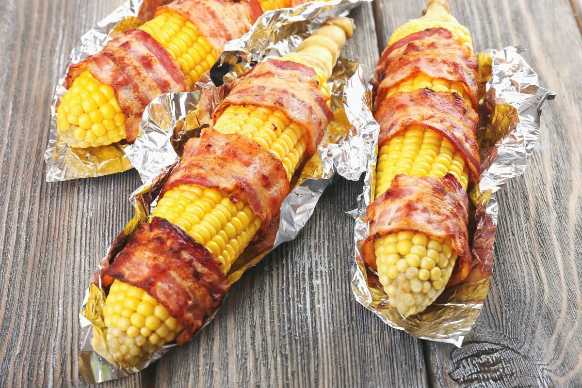 3 Bacon Wrapped Corn on the Cobs sitting on foil on a rustic surface.