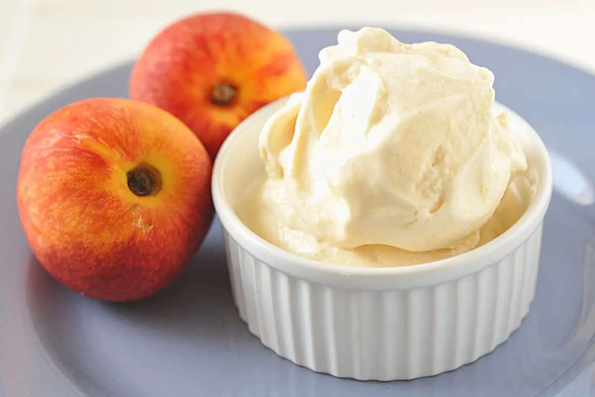 A scoop of Homemade Peach Ice Cream in a white dish with 2 fresh peaches next to it.