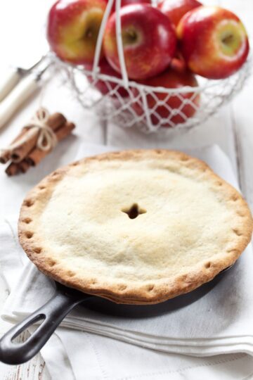 Easy Skillet Apple Pie on a white table cloth with a basket of apples, and a bunch of cinnamon sticks behind it.