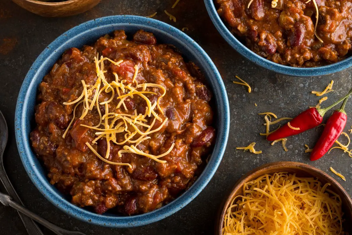 A hearty bowl of chili topped with shredded cheese, served in a rustic blue bowl with a side of shredded cheese and red chili peppers on a dark surface.