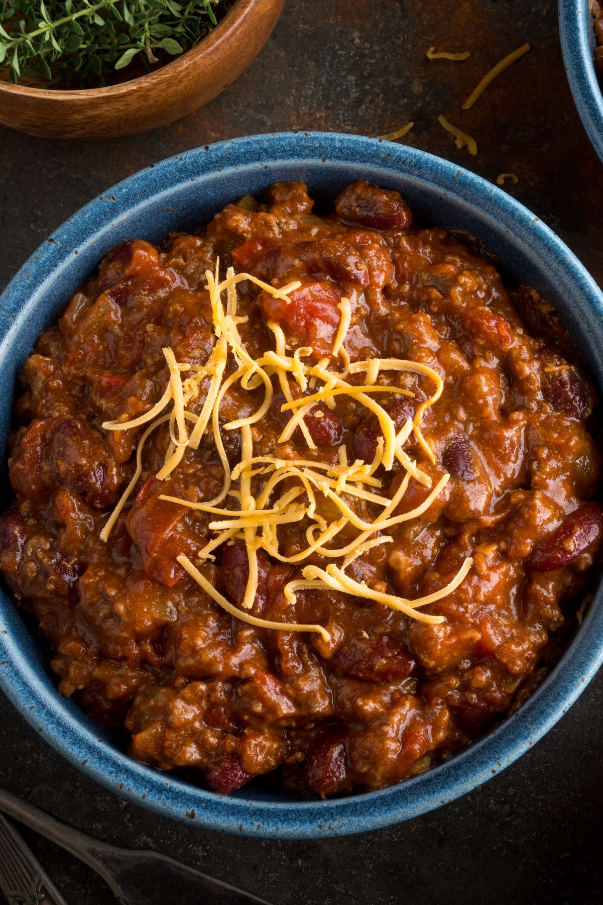 Close-up of a bowl of rich and thick chili garnished with shredded cheese, in a blue bowl against a dark, textured background.