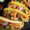 Photo of crunchy yellow corn tacos filled with shredded Slow Cooker Mexican Chicken Taco Meat, topped with fresh radishes, cucumber slices, and a creamy sauce. They are served on a black plate accompanied by lime wedges, showcasing a colorful and appetizing meal option.