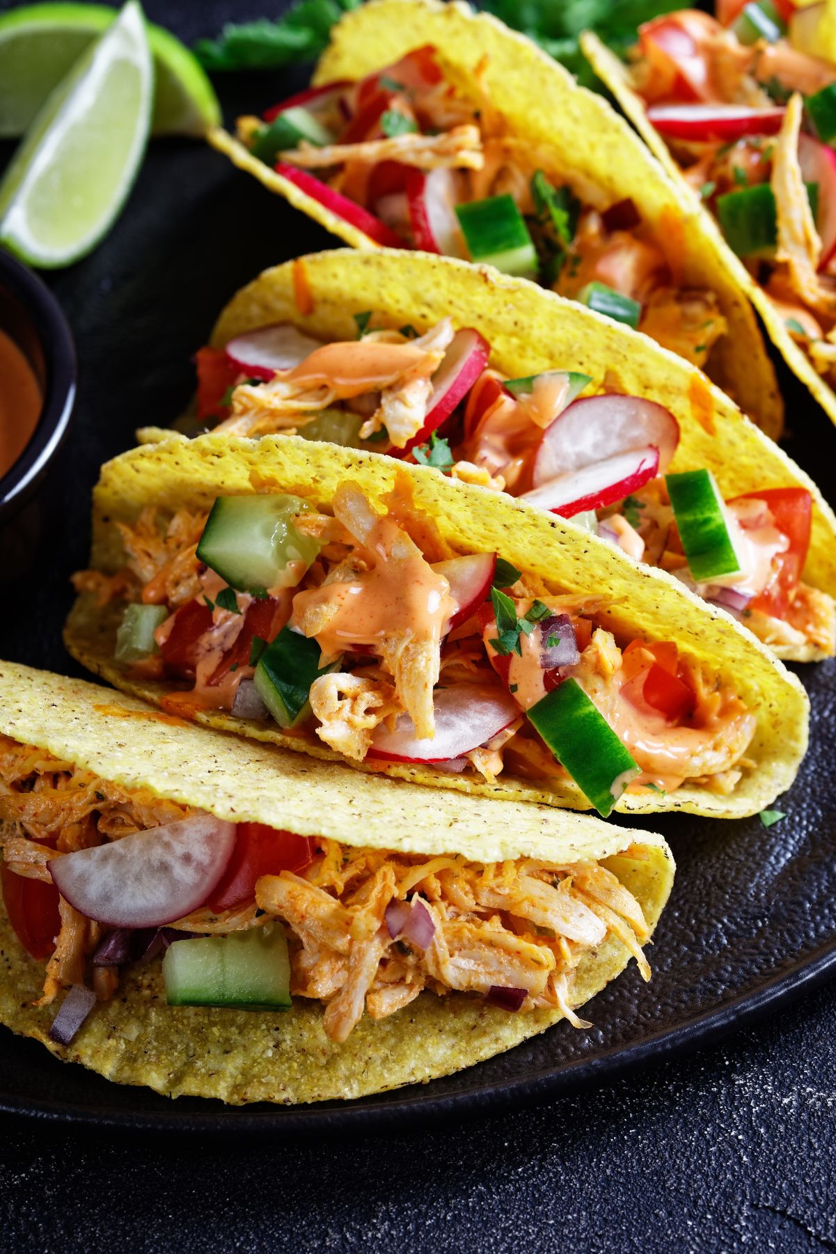 Photo of crunchy yellow corn tacos filled with shredded Slow Cooker Mexican Chicken Taco Meat, topped with fresh radishes, cucumber slices, and a creamy sauce. They are served on a black plate accompanied by lime wedges, showcasing a colorful and appetizing meal option.
