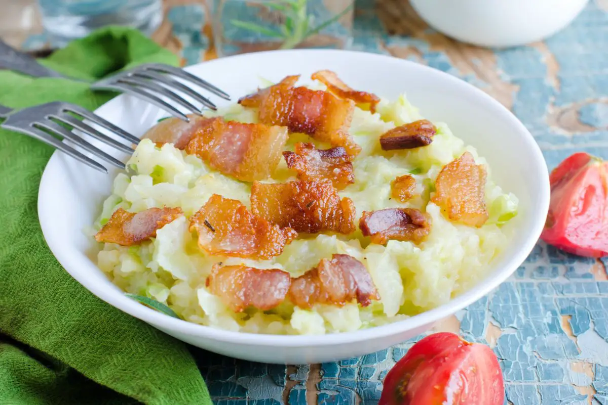 A bowl of Colcannon, featuring creamy mashed potatoes mixed with bits of green cabbage, topped with crispy, golden-brown bacon pieces, presented on a blue rustic table alongside a green cloth napkin and a fresh red tomato.