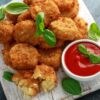 Crispy fried macaroni and cheese balls are arranged on a white, rustic wooden paddle board, with a few basil leaves for garnish. One ball is broken open, showing the gooey cheese inside. A small white bowl with tomato sauce is also on the board, ready for dipping.