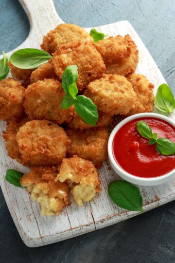 Crispy fried macaroni and cheese balls are arranged on a white, rustic wooden paddle board, with a few basil leaves for garnish. One ball is broken open, showing the gooey cheese inside. A small white bowl with tomato sauce is also on the board, ready for dipping.
