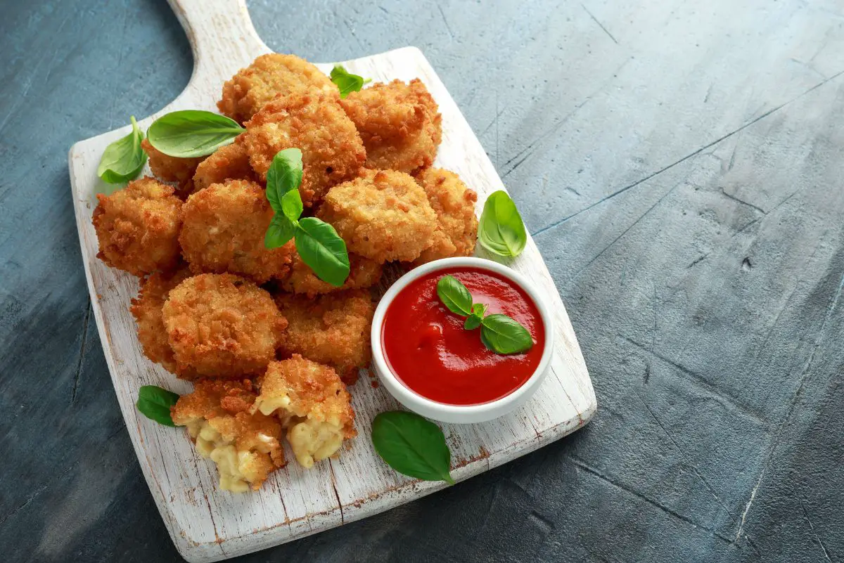 A wooden serving board holds golden-brown fried macaroni and cheese balls, garnished with fresh basil leaves, alongside a small bowl of red dipping sauce, on a dark blue textured surface.