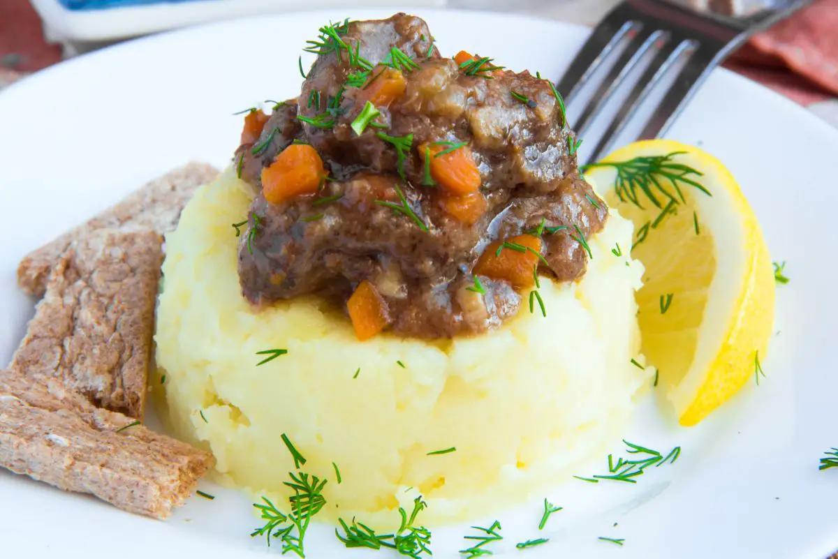A plate of Irish stew served on a bed of mashed potatoes garnished with dill, accompanied by a lemon wedge and slices of brown bread.