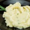 A serving of Irish Champ, featuring creamy mashed potatoes with flecks of green onion, on a dark plate set on a wooden table.