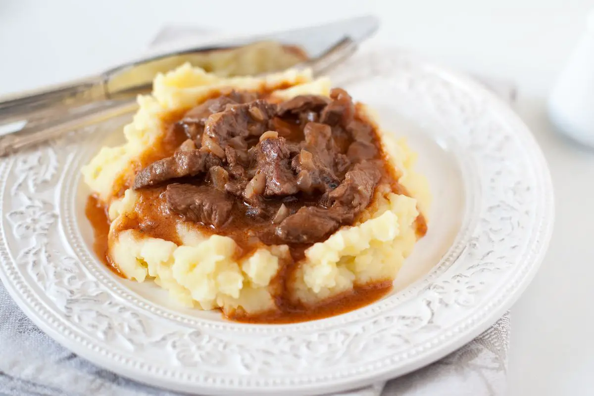 A serving of Irish Guinness beef stew, with chunks of beef in a rich, brown sauce, served over a bed of creamy mashed potatoes on an ornate white plate.