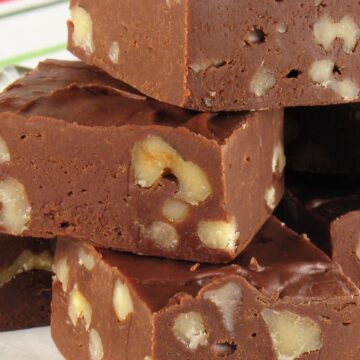 A close-up view of square-cut pieces of chocolate fudge filled with whole nuts, stacked on a white decorative plate. The fudge has a dense and smooth texture, with the nuts evenly distributed throughout, and the plate sits on a surface with a red and green striped napkin, adding a holiday feel to the image.