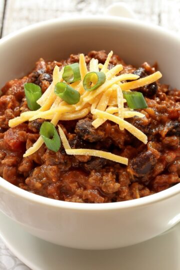 A close-up view of a bowl of The Ultimate Homemade Chili, topped with shredded cheddar cheese and green onion slices, on a rustic wooden table background.