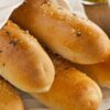 A detailed close-up of oven-fresh, soft breadsticks with a shiny, golden crust, topped with fine granules of salt and specks of green herbs, arranged on a white cloth over a rich, dark wooden table, with a soft-focus view of a glass container of olive oil to the side.